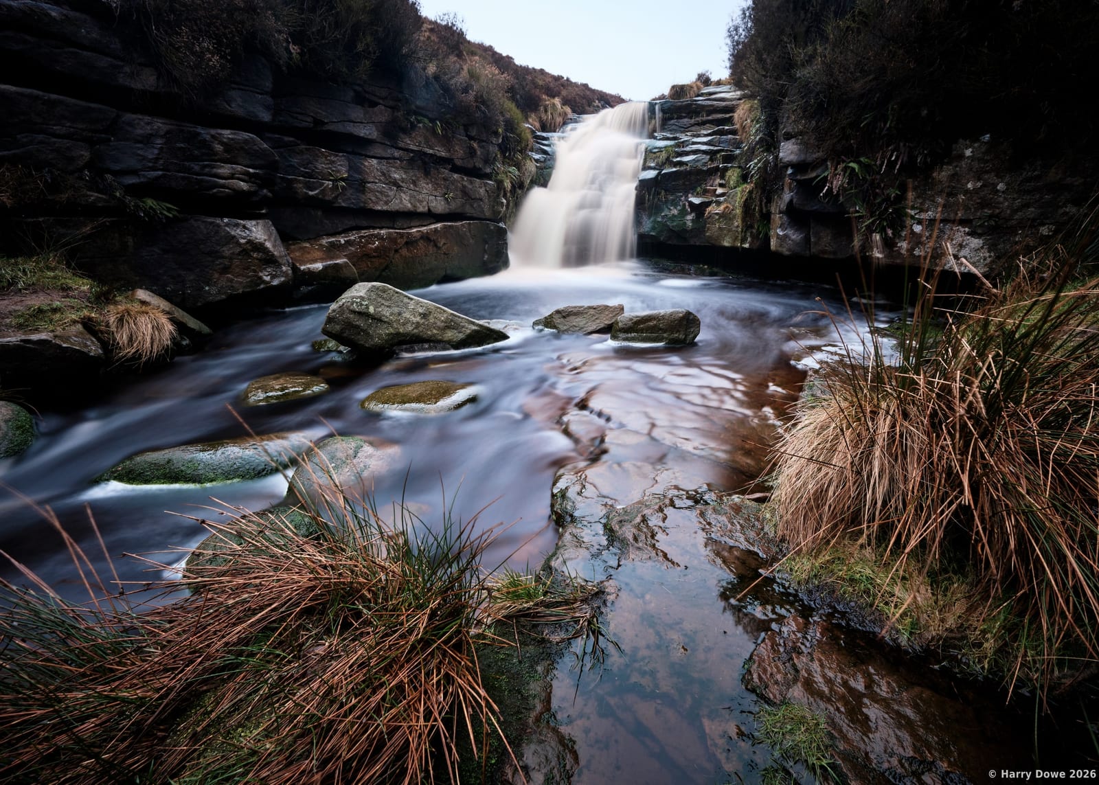 Long exposure photograph showing silky water cascading over dark rock steps into a clear pool surrounded by moorland grasses
