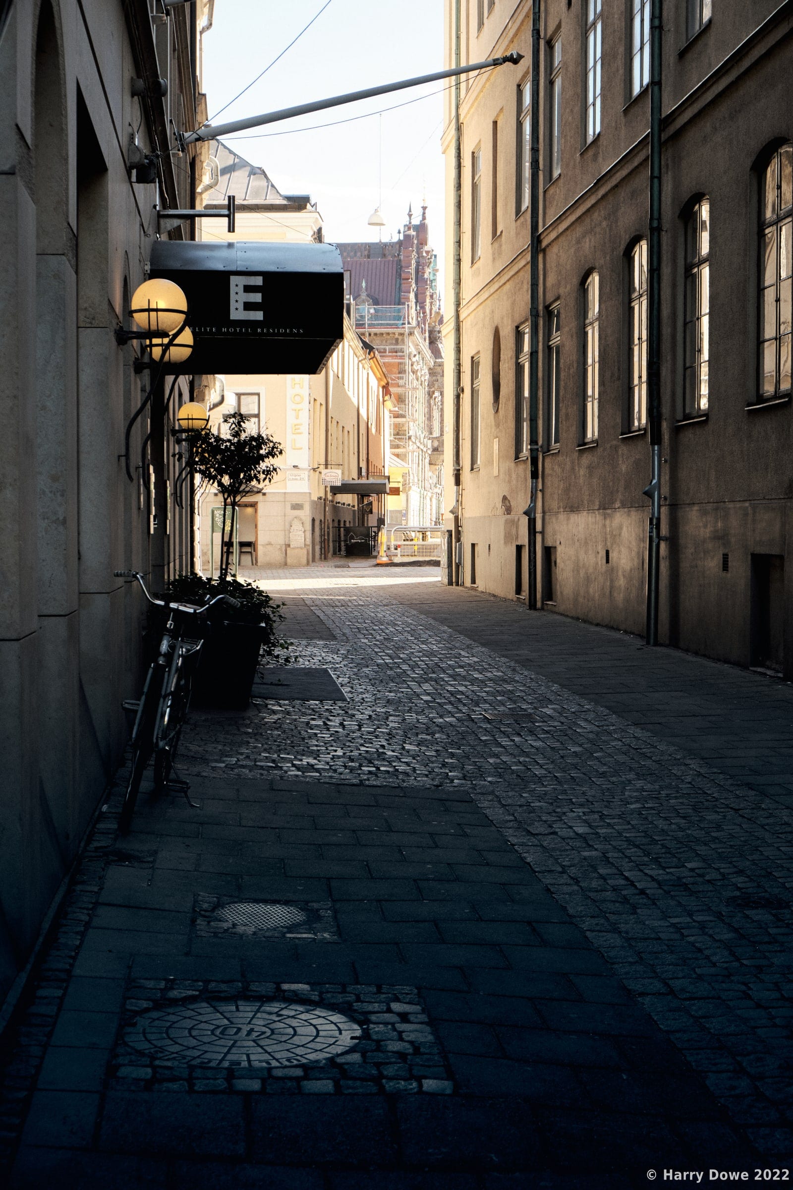 Narrow cobblestone European alleyway with tall historic buildings, glowing globe wall lamps, and a bicycle parked in the foreground shading into bright sunlight ahead