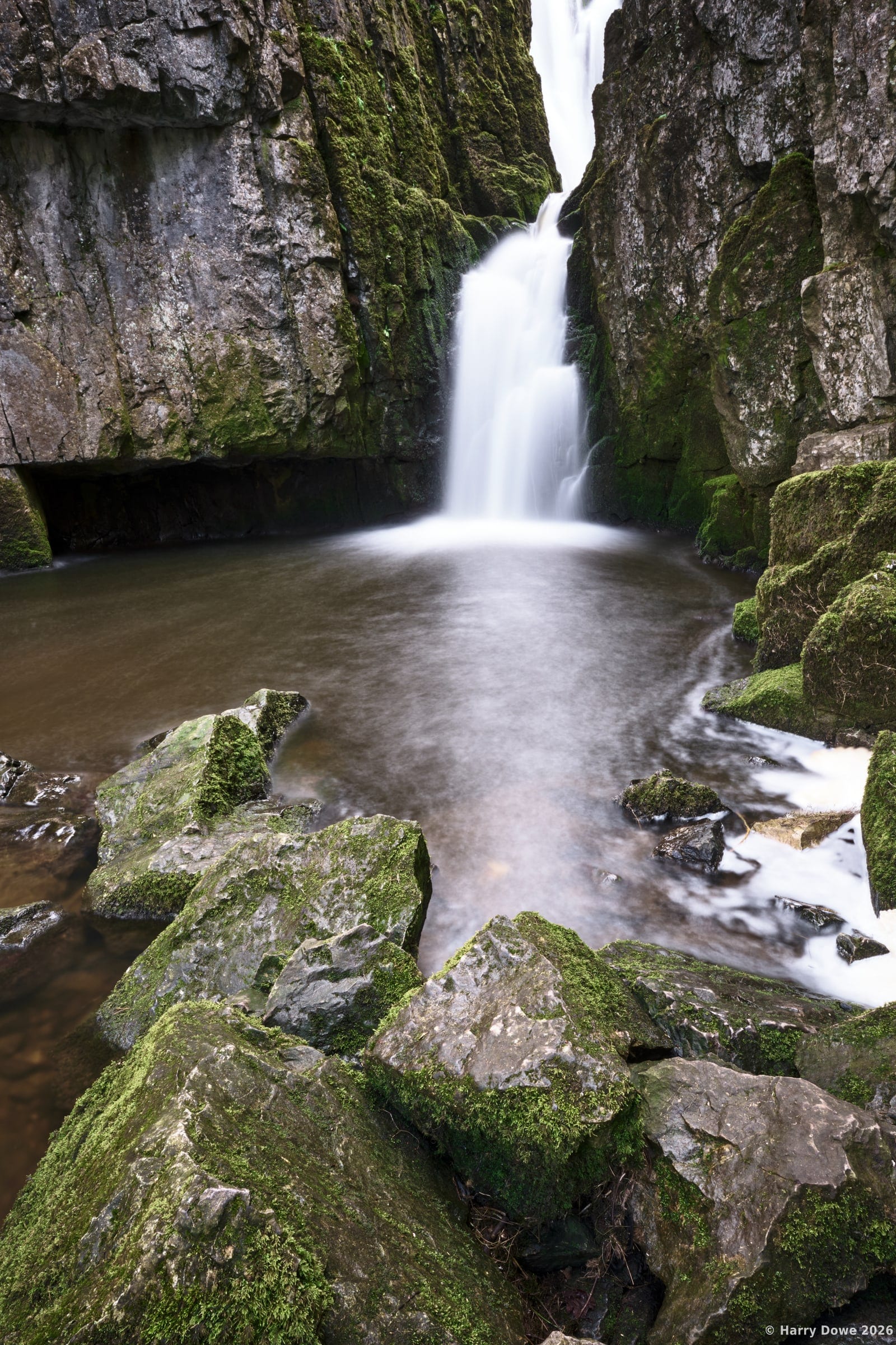 Vertical long exposure shot of a powerful waterfall plunging between steep mossy rock walls into a misty pool surrounded by lichen-covered stones