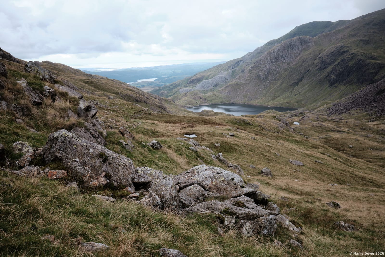 Wide vista of rocky, grassy mountain slopes looking down toward a quiet lake nestled in a sweeping valley under a cloudy sky
