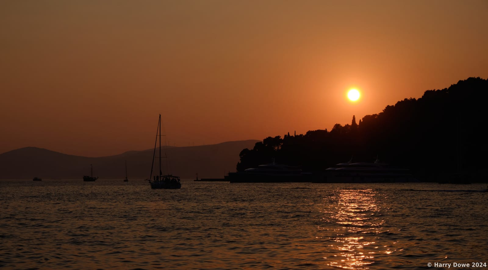 Silhouetted sailboat navigating calm coastal waters during a fiery amber sunset with light reflecting off the ripples and hills in the background