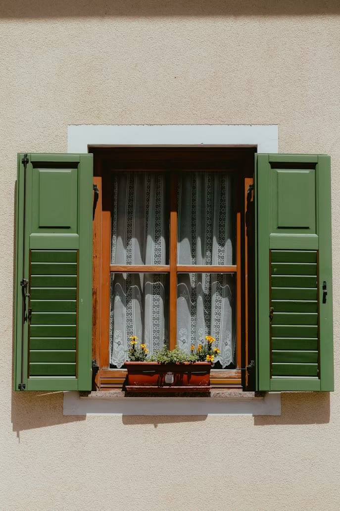 Green shutters framing a lace-curtained window with a flower box on a sunlit cream wall.