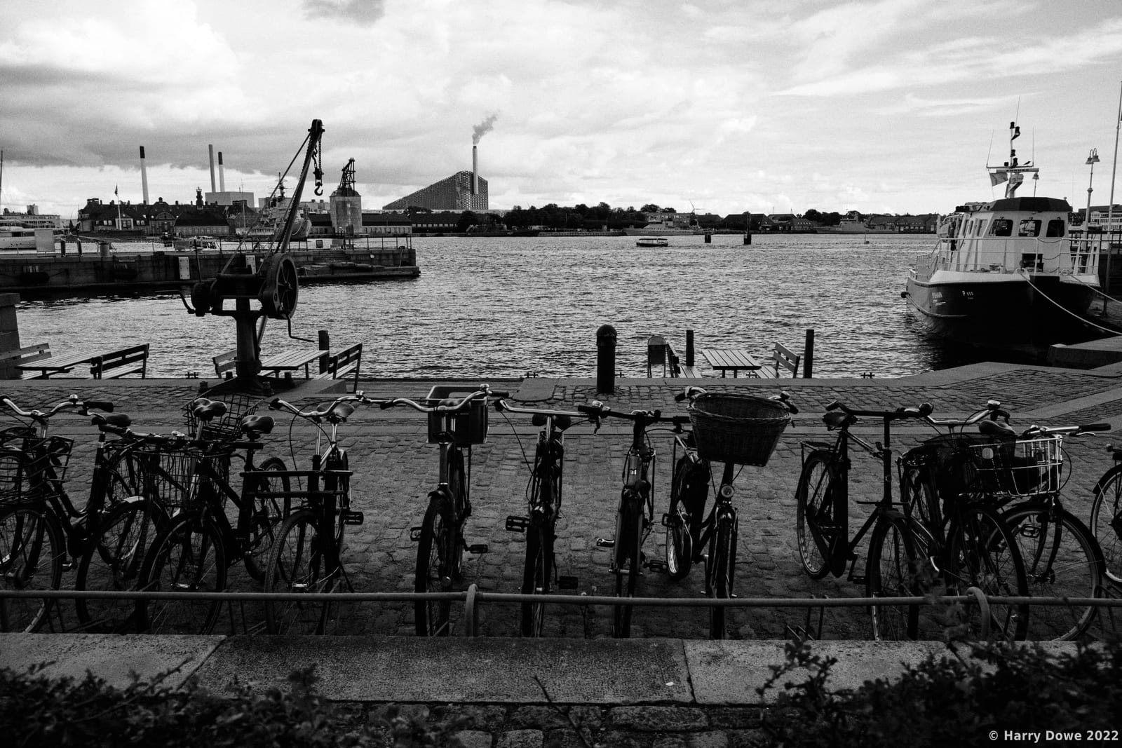 Black and white high-contrast photograph of bicycles parked along a stone harbor promenade with boats, cranes, and industrial skyline across the water