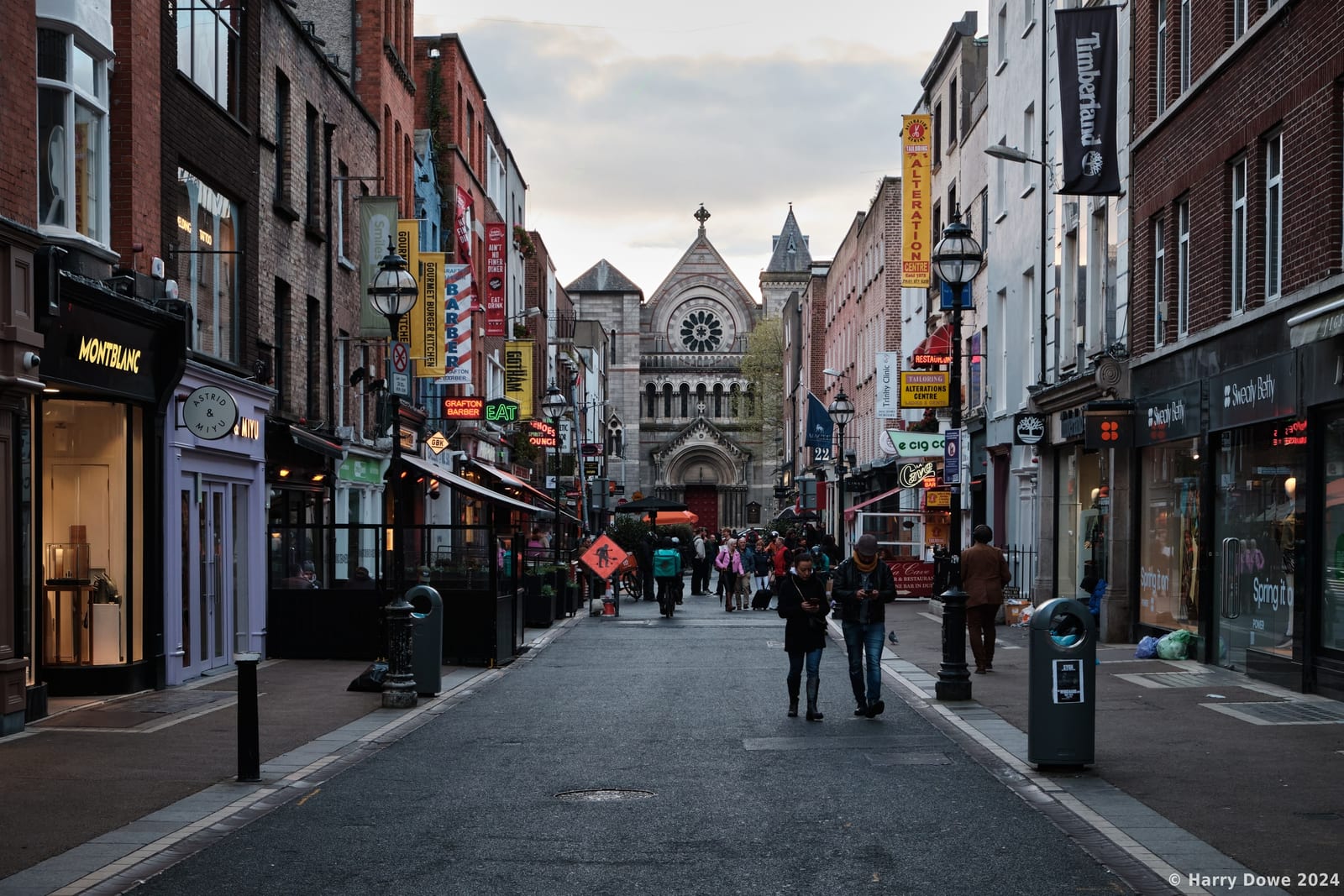 Pedestrians walking down a lively urban shopping street lined with shops and hanging storefront signs leading toward a large stone gothic church at the end of the vista