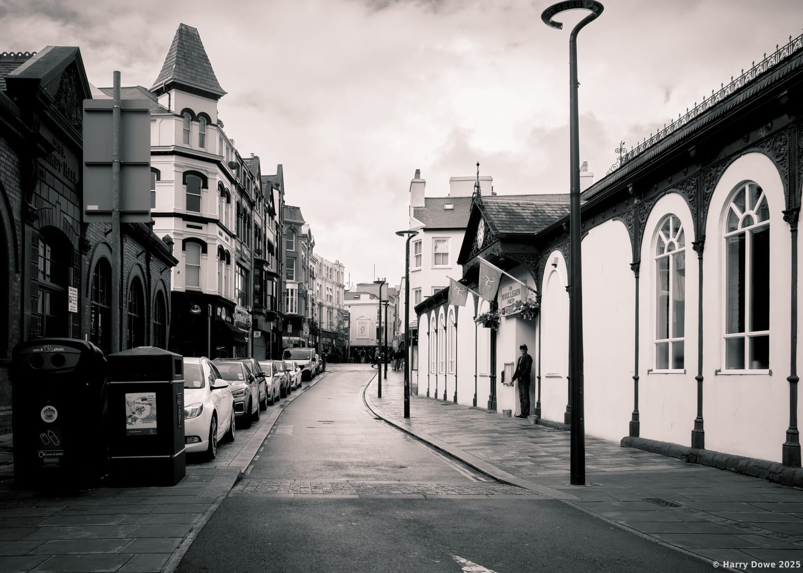 Moody monochrome perspective of Victoria Street in Douglas showing classic Manx architecture, arched windows, and parked cars stretching into the distance.