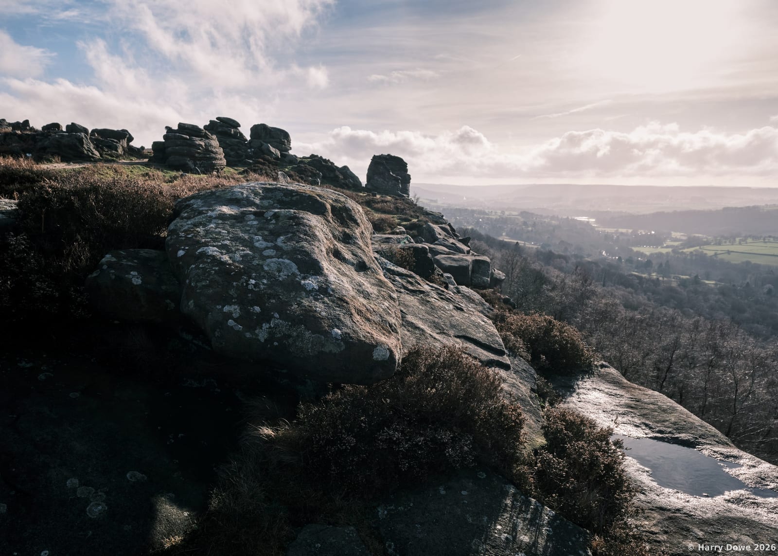 Large moss-covered boulder and gritstone edge bathed in soft sunlight, overlooking a misty, vast valley floor below.