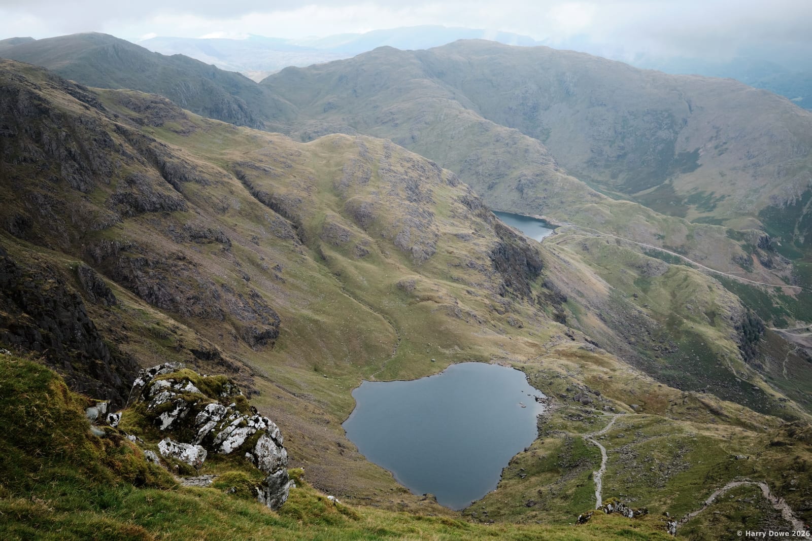 High elevation perspective looking down on a dark mountain tarn and winding trails nestled among massive, rolling mountain ridges under an overcast sky