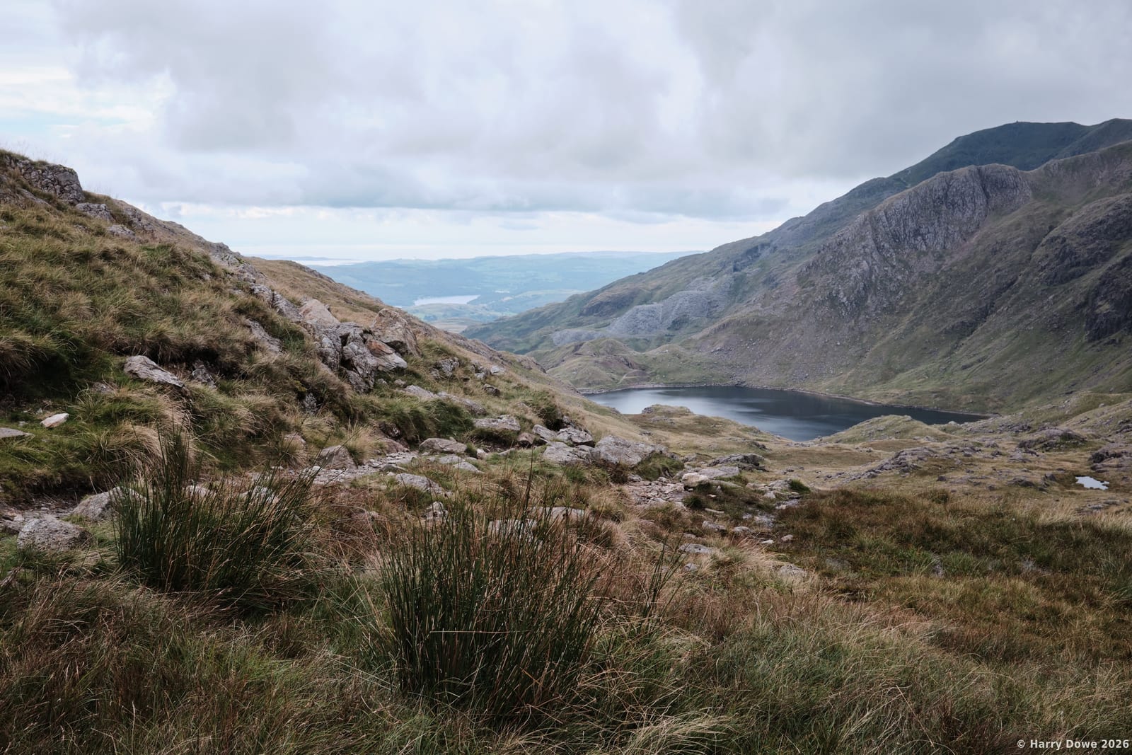 High angle view looking down grassy slopes toward a dark mountain tarn cradled between steep rocky ridges beneath an overcast sky