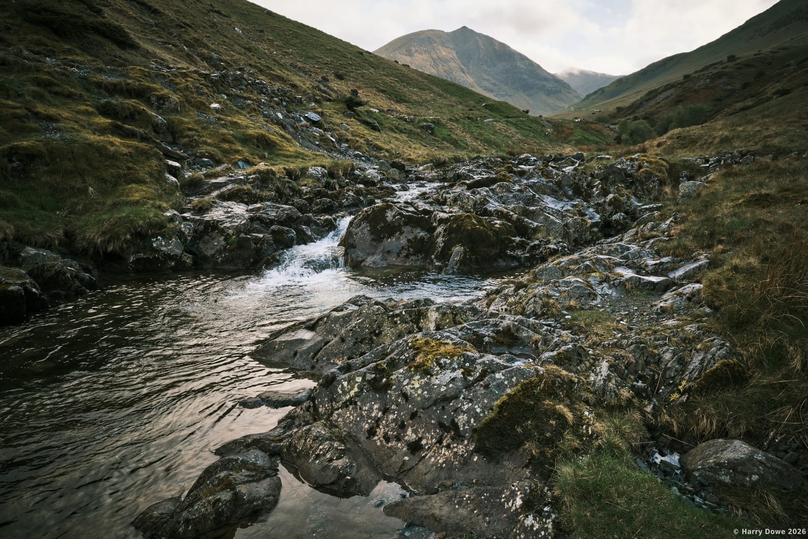 Rocky mountain stream winding through a steep green valley with majestic peaks looming in the atmospheric distance