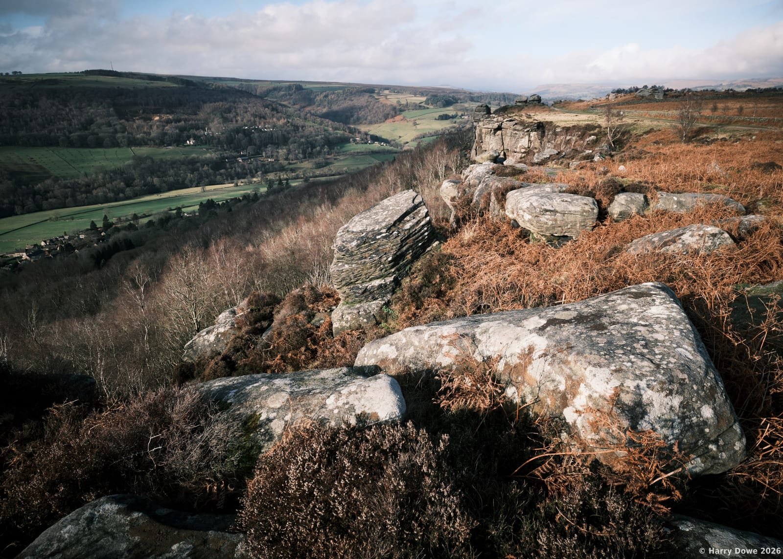 Weathered boulders and tawny bracken lining a high ridge with a sprawling green valley and distant countryside below.