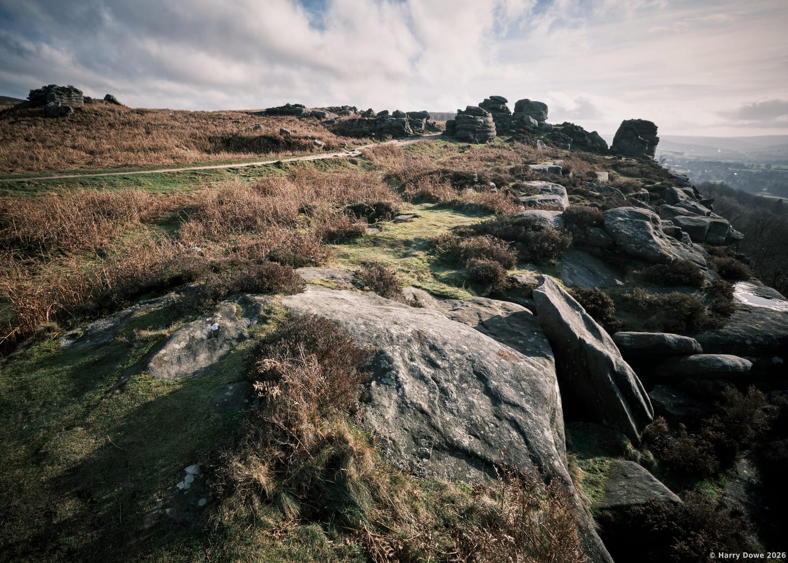 Weathered gritstone rock formations jutting out from wild moorland heather along a windswept ridge overlooking the valley below
