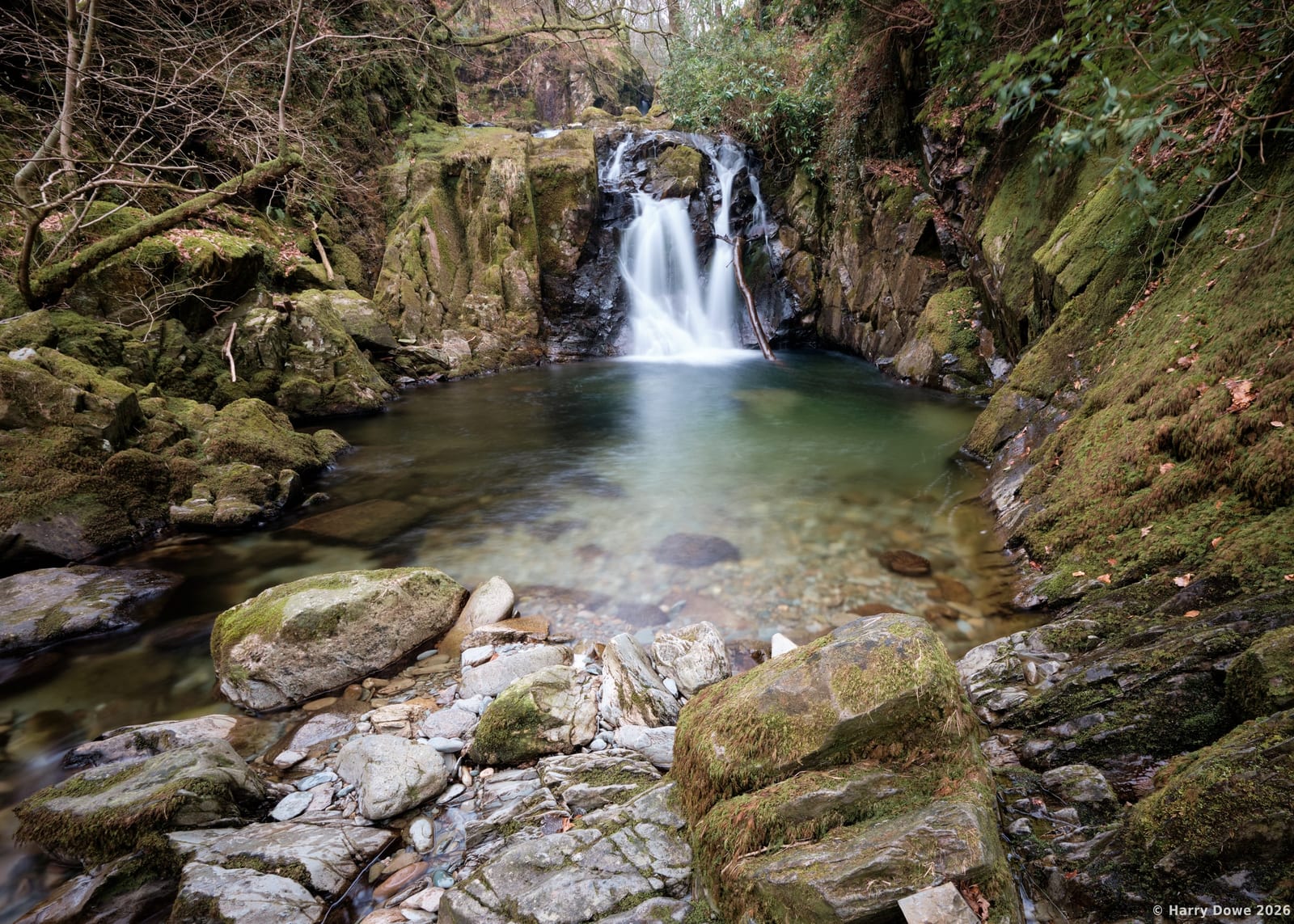 Silky waterfall tumbling over dark rocky cliffs into a clear, tranquil forest pool surrounded by mossy rocks and lush trees