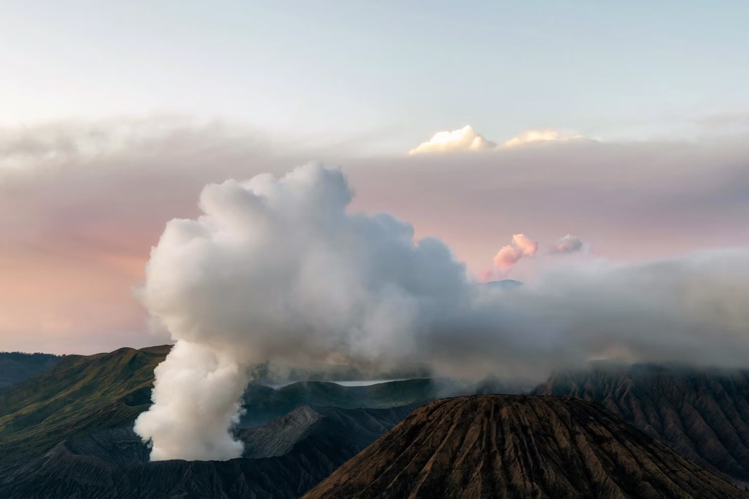 A plume of white steam rising from a volcanic crater through low clouds at sunrise.