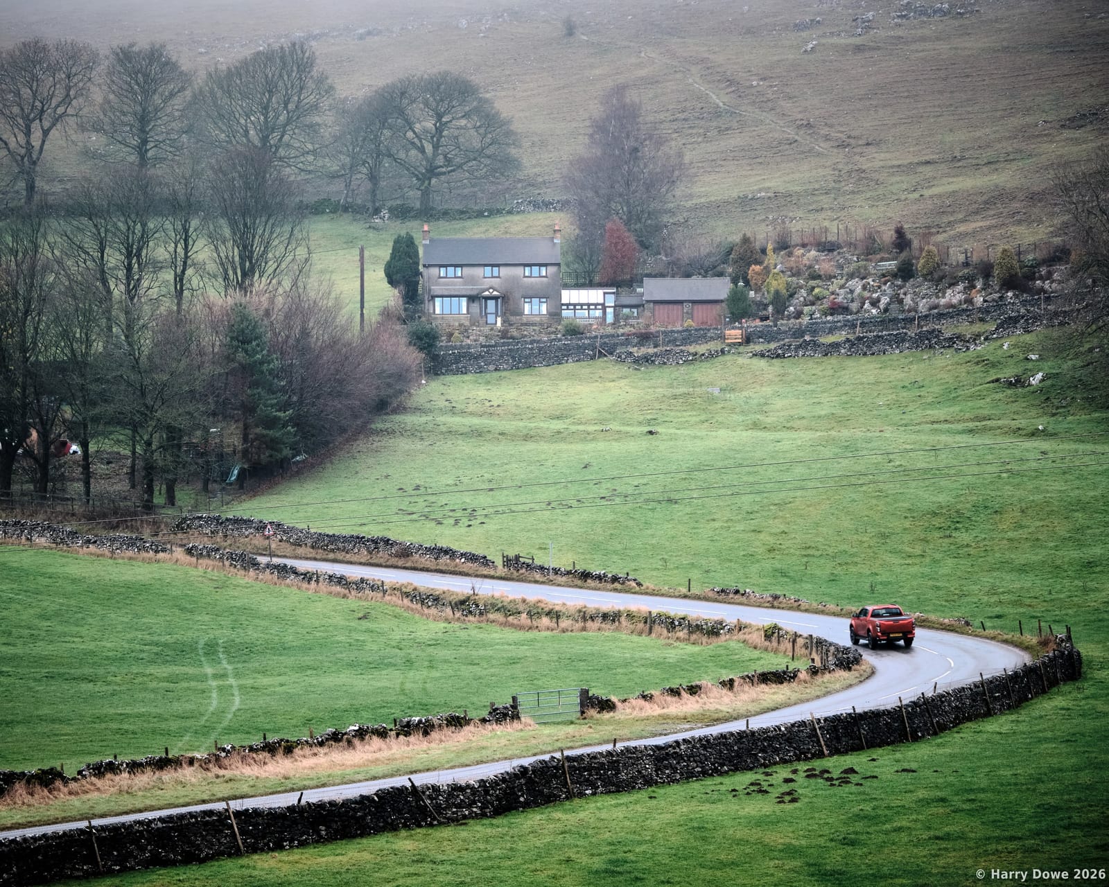 A red pickup truck driving along a curving asphalt road lined with stone walls, sweeping past rolling green fields toward a remote hillside cottage
